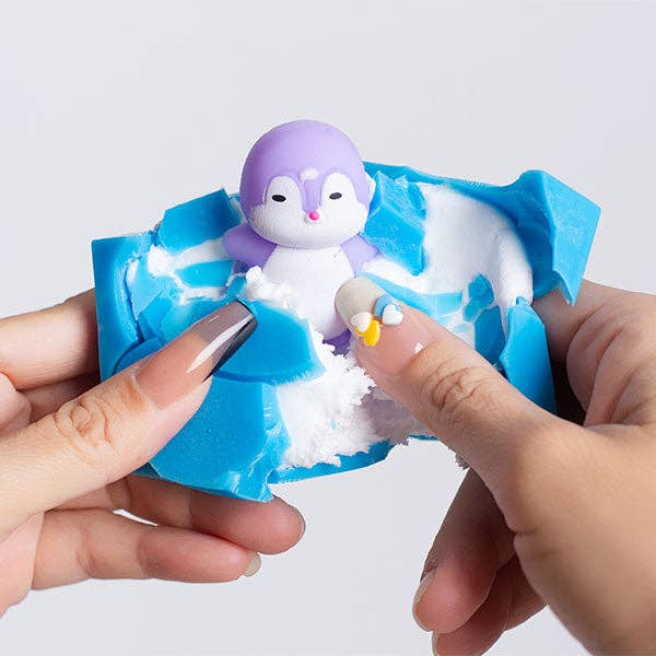 A small penguin figurine with a purple head and white body nestled in torn blue and white material, held between two hands against a light gray background.