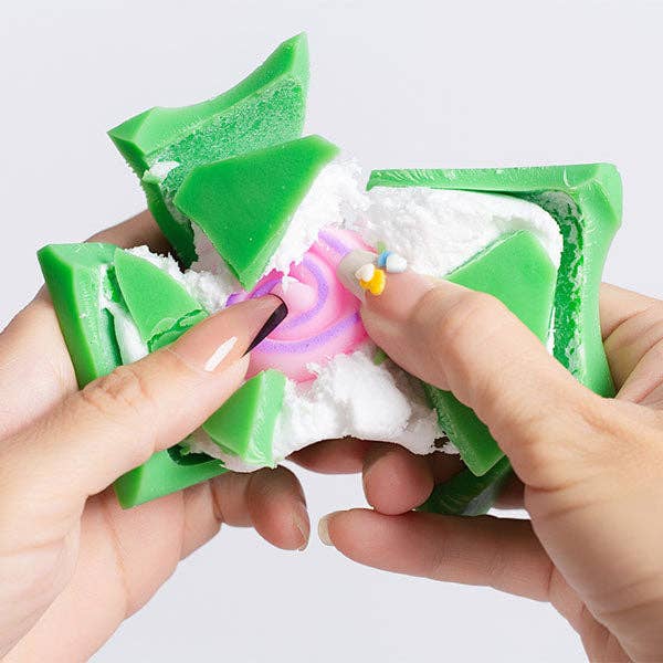 A close-up of two hands holding and crushing a green and white squishy toy with a pink and purple swirl center, against a light gray background.