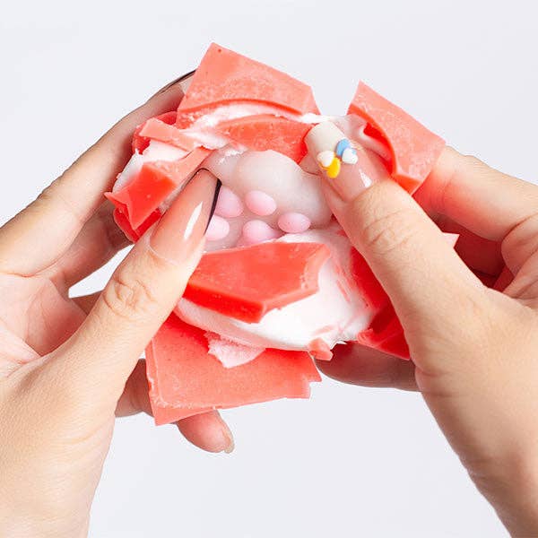 A close-up of hands peeling back translucent pink and white wax-like sheets, revealing small pale pink beads underneath, against a plain light background.