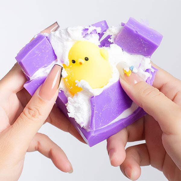 Hands cracking open a purple Wax Clay Squishies blind box to reveal a yellow baby chick squishy toy inside.