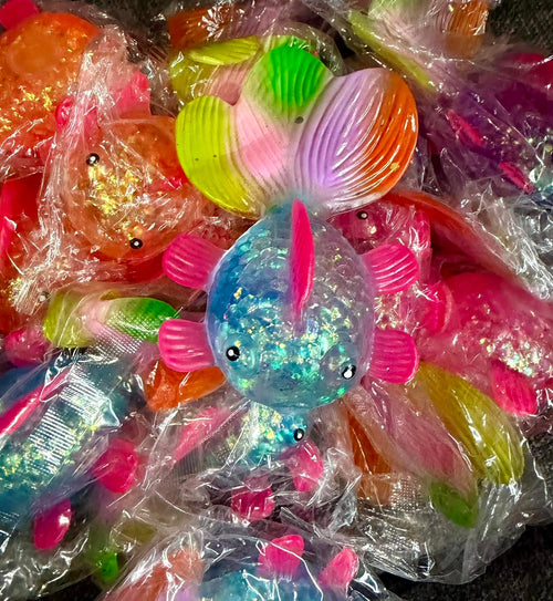 A pile of colorful, glittery fish-shaped squishies wrapped in clear plastic bags. The central fish is blue with pink fins and a multicolor tail, surrounded by other similarly packaged fish in various colors.